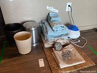 Photo of small trash containers, bath linens, bamboo tile mat, humidifier, shower head, and shower hooks on floor.