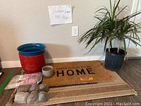 Photo showing area rugs including 'Feels Good to Be Home' mat, living plant in pot, red and blue planters, small gray pot, outdoor paving stones, packaged gardening gloves, wooden plant stakes labeled 'Outdoor/Paper Pots/Rogs'.