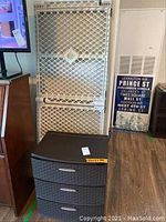 Front view showing folded heavy plastic baby playpen and black three-drawer cabinet with woven drawer fronts.
