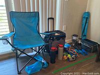 Photo showing two blue folding camping chairs with black metal frames, cooler, water bottles and assorted camping gear on floor near a window.