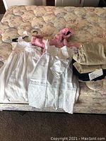 Wide view showing all items together: two white christening dresses, pink hooded jacket, and three pairs of folded school uniform pants.