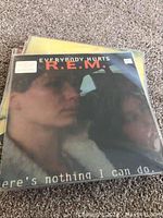 Photo of vinyl record album cover R.E.M. titled "Everybody Hurts" showing man and woman in car, with visible protective plastic covering.