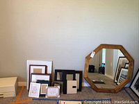 Photo shows large octagonal wood framed mirror next to multiple empty picture frames of various shapes and sizes arranged against a wall on a carpeted floor.