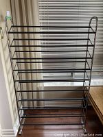 Front view of standing shoe rack showing 10 black horizontal bars supported by chrome frame with rounded handles on sides.