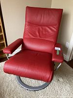 Front three-quarter view showing red leather upholstery, high backrest, padded armrests and round brushed nickel base