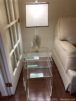 Three nesting tables stacked, with lamp on top next to a beige upholstered chair and white door.