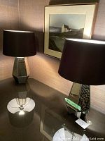 Pair of black mirrored lamps with hexagonal bases and black fabric shades, shown on a reflective surface with framed wall art in the background.