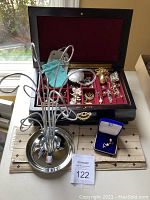 Dark wood jewellery box open to reveal red velvet interior, assorted pins, and costume jewellery inside; chrome jewellery tree is positioned next to the box; jewelry organizer bags partially visible; Nash Jewellers earring box with earrings in front