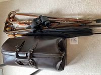 Photo showing vintage brown leather bag, 5 wooden canes stacked on top, black umbrellas, and Bushnell binoculars placed on beige carpet.