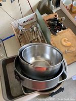Photo showing stainless mixing bowls stacked, metal pans including springform, 9x9 and 9x12 cake pans, and cutlery rack with spoons and forks.