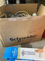 Photo showing a cardboard box labeled 'Schneider Electric' filled with assorted cords, bulbs, and the corner of Aqua Sense grab bars in packaging below it.