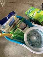 Mop bucket with wringer containing cleaning brushes, sponges, and sealed packets, two boxes of new Swiffer Sweeper refills nearby