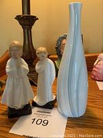 Front view of two Royal Doulton figurines and a tall white vase on a wooden surface.