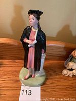 Front view of Royal Doulton figurine The Graduate in gown and cap holding diploma, standing on light green base.