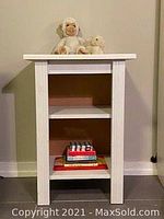 Front view of the small white wooden bedside table showing two shelves, pink back panel, and items placed for display.