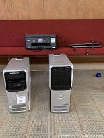 Two Dell Dimension desktop towers placed upright on carpeted floor in front of a red bench, showing front panels with drives and power buttons. One is Dimension E520 and the other Dimension 9200 model.