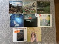 Photo showing eight vintage vinyl record albums arranged on a carpet. Albums vary in genre including orchestras, choirs, nature sounds, and vocal performances. Covers feature scenic photography, illustrated animals, and performer portraits.