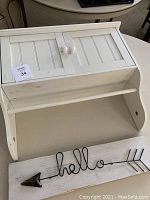 White wooden hanging cabinet with two doors and dowel showing some staining and missing dowel; Hello wooden sign with wire 'hello' text and arrow mounted on a white base with some chipping at bottom