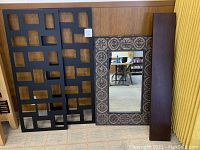 Two large wall frames with multiple square openings, decorative framed mirror with patterned edges, and dark wood shelf stacked together leaning on a wood paneled wall