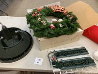 Photo showing artificial Christmas wreath with small ornaments and red-gold bow, black metal tree stand, and segments of string lights on a table.