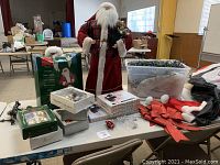 Photo showing large Santa Claus figure, Santa suit pieces, assorted Christmas decorations including ornaments, bows, lights, and hats on a table.