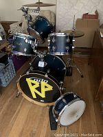 Full drumset shown with bass drum featuring black head with yellow RP decal, and visible tom toms with blue gradient finish.