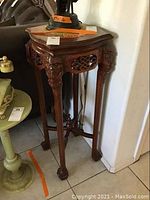 Full view of tall wooden pedestal with intricate carved details on legs and apron near corner wall and green round table next to it