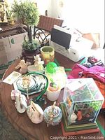 Wide assortment of decorative items including porcelain figurines, green curved glass vase, small flower pot, a colorful music box with glass enclosure containing artificial flowers and seashells, all arranged on a wooden table.