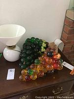 Photo of two colored glass grape bundles and white mid century table lamp on a wooden surface.