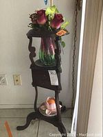 Front view of dark wood three-tiered stand holding glass vase with artificial flowers on top, two drawers in middle, and ceramic rose dish with orange ball at bottom.