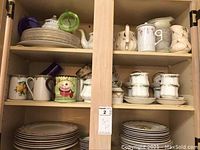 Shelf showing stacked plates, several teapots, and assorted teacups including a colorful cupcake mug.