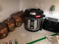 Countertop view of pressure cooker, four ceramic canisters with apple designs, and black toaster