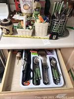 Image of flatware organized in a drawer with green handles on forks, knives, spoons, and more, alongside three ceramic canisters on countertop.