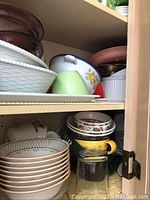 Ceramic dishes and bowls with floral and geometric patterns, white bowls with gold trim, clear glassware with green and polka dot lids in a cabinet shelf.