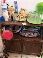 Wooden cabinet containing ceramic serving set, glassware, glass bottles, plastic tray, and kitchen decor on and inside shelves