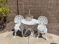 Full view of white wrought iron bistro table with two chairs and the planter on table, set outdoors on stone and concrete.