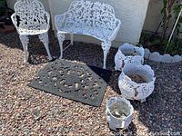 Photo showing the white wrought iron chair, bench, three planters, and black outdoor mat on gravel surface outside against a wall.