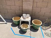 View showing three green ceramic pots and three small white ceramic pots arranged on and around white plastic table outdoors.