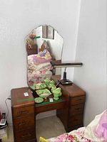Photo showing antique wooden desk with attached mirror, green ceramic items (not included), dark base lamp on top, and floral bedding adjacent.