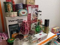 Overview of the lot showing Hamilton Beach coffee maker box, decorative candles, vases, and decorative tin boxes clustered on a table
