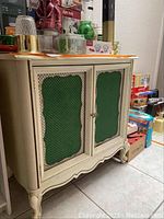 Front view of white antique cabinet with green fabric under metal mesh doors and decorative wooden trim.