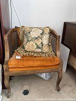 Front view of antique wooden chair with wicker panels on sides, orange cushioned seat and back, floral fringed throw pillow included