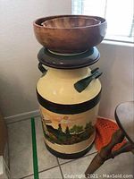 Painted metal milk can with black bands and colorful countryside scene showing trees and sun, topped with a larger wooden bowl and a smaller scalloped wooden bowl inside