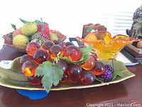 Photo showing resin grapes on green ceramic platter, faux fruit bowl, orange glass vase, and wooden napkin holder on table