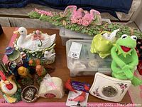 Display of assorted collectible figurines including ceramic duck planter, colorful bird figurines, frog planter, Kermit the Frog plush, ceramic clock with floral motifs, and other miniature decorative items on wooden table.