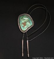 Full view of the bolo tie showing the large turquoise stone in an irregular shape set in silver bezel mounted on braided leather tie with silver aiguillettes at the end.