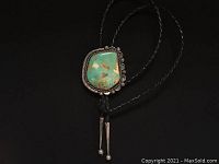 Full view of the bolo tie showing the green turquoise stone set in sterling silver bezel with braided black leather tie and silver aiguillettes.