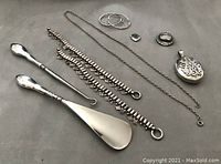 Overhead photo showing the shoehorn, boot hook, two bracelets with some missing dangle discs, pair of silver hoop earrings, heart locket, brooch, and large hanging locket with chain arranged on a flat surface.