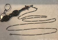 Full view of the lorgnette with handle and attached long chain with clear cabochons placed on a neutral background.