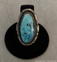 Front view of a sterling silver ring showcasing a large oval turquoise stone surrounded by a handcrafted silver halo with detailed edge work on a black ring stand.